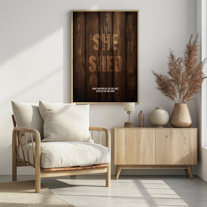 A rustic poster with a dark brown wood panel background. The words 'SHE SHED' are in large, blocky letters with a distressed wood texture. Below, a smaller caption reads 'WHAT HAPPENS IN THE SHE SHED STAYS IN THE SHE SHED'. The poster is displayed in a thin black frame. Print