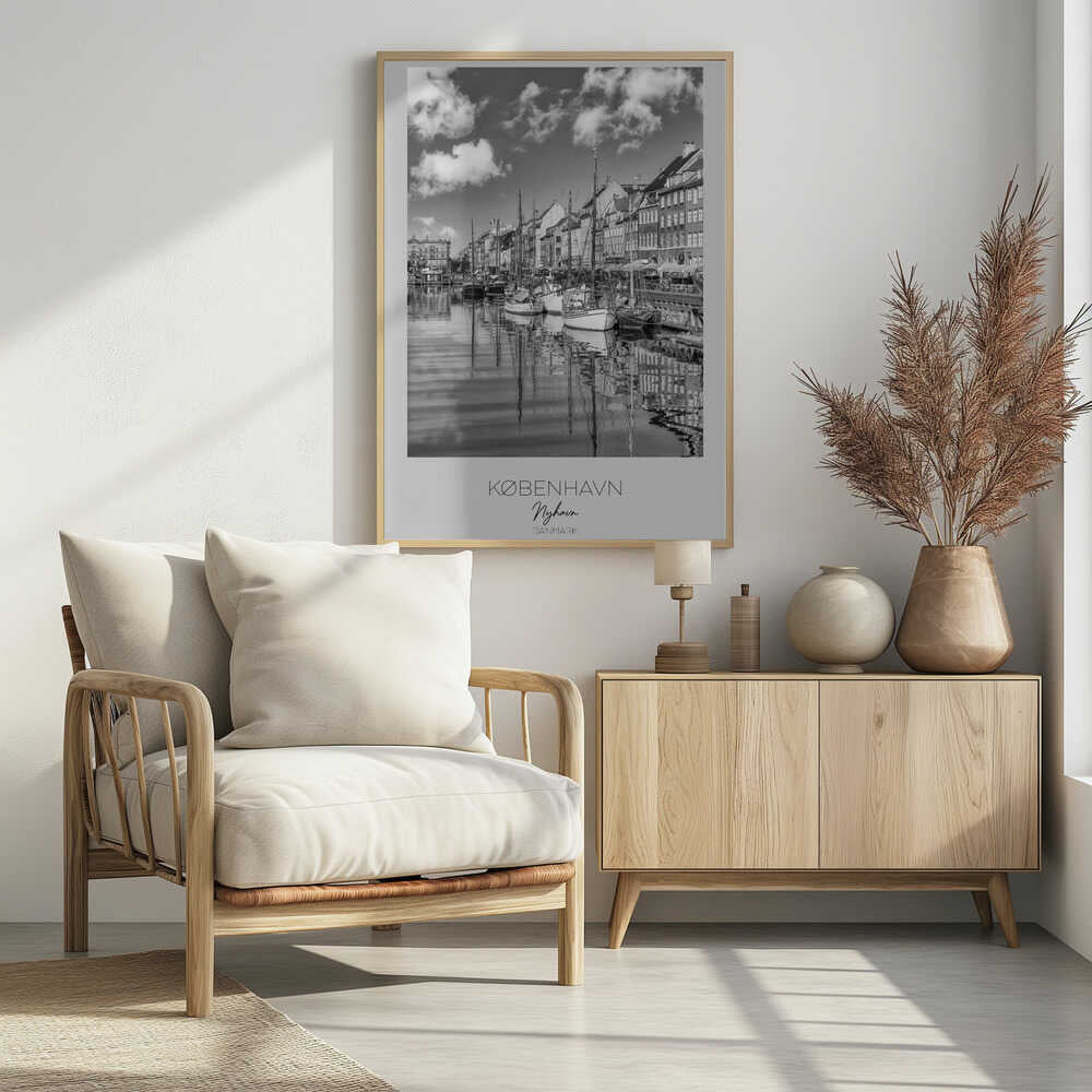A black and white poster of Nyhavn harbor in Copenhagen. The photograph shows traditional sailboats and ships moored along a canal, with a line of historic, multi-story buildings on the far bank. The sky has fluffy clouds, and the entire scene is reflected in the calm water. The poster has a white border with the text 'KØBENHAVN', 'Nyhavn', and 'DANMARK' at the bottom, and is shown within a black frame. Poster