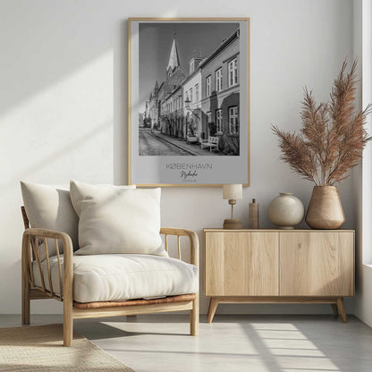 A black and white framed travel poster of a charming cobblestone street in Nyboder, Copenhagen. The photograph shows a sunlit row of historic houses with a prominent church steeple in the background. At the bottom of the poster, the words 'KØBENHAVN', 'Nyboder', and 'DANMARK' are written. Wall Art