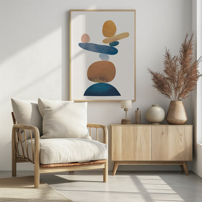 An abstract art print in a thin black frame featuring a stack of colorful, organic shapes resembling balanced stones against a white background. The forms are in various shades of blue, earthy brown, ochre, and a muted pink, creating a sense of calm and equilibrium. Artwork