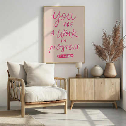 A framed art print featuring the motivational quote 'You are a work in progress'. The text is rendered in a vibrant pink, hand-lettered style against a soft beige background. The words 'You', 'a Work', and 'progress' are in a flowing script, while 'are' and 'in' are in a simple uppercase font. Wall Art