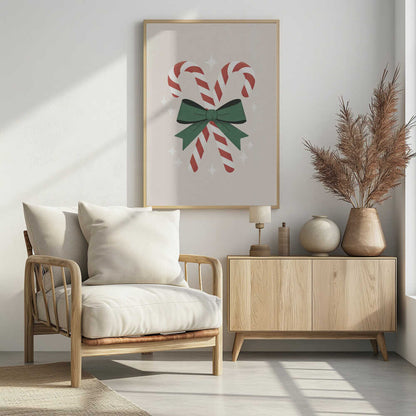 An illustration of two red-and-white striped candy canes crossed and tied together with a large green bow. The background is a soft beige color with small, white, four-pointed star sparkles. The artwork is shown in a black frame. Decor