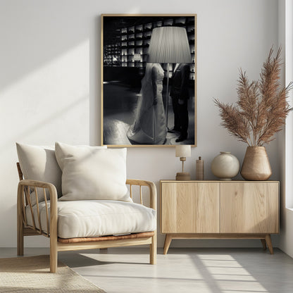 A surreal black and white photograph of a bride and groom whose heads are comically obscured by a large, pleated lampshade from a floor lamp standing between them. They are in a modern, dimly lit room with a wall of illuminated shelves. Poster