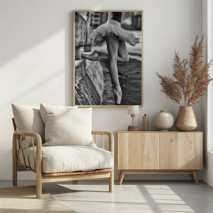 A black and white photograph focusing on the lower half of a ballerina in a tutu and pointe shoes. She stands en pointe with one foot on the ground, while the other foot rests against a rough, concrete curb, highlighting the contrast between delicate dance and an urban environment. Decor