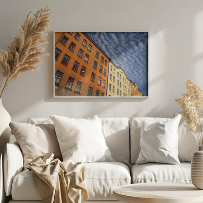 A low-angle shot of colorful buildings, predominantly a vibrant orange one, against a bright blue sky filled with a pattern of white, fluffy clouds. The image is presented within a silver-colored frame. Print