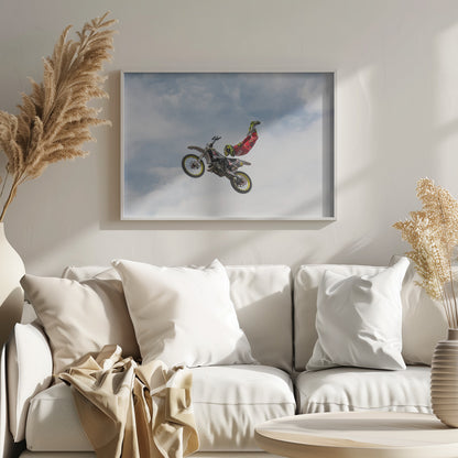A motocross rider in red and yellow gear performs a high-flying stunt, arching their back while upside down in the air next to their dirt bike, set against a backdrop of a blue sky with white clouds and enclosed in a silver frame. Poster