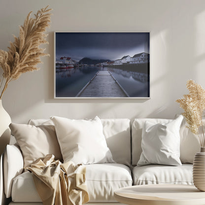 A long exposure photograph of a serene coastal town at night, viewed from a wooden pier that leads into calm, dark water. A lone person stands at the end of the pier, gazing at the town's reflected lights. Behind the village, a mountain looms under a breathtaking, star-filled sky. The image is presented within a silver-colored frame. Artwork