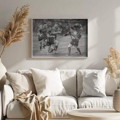 A dynamic black and white photograph of a rugby match being played in the rain. One player charges forward with the ball while an opponent wraps his arms around him in a tackle. Other players are in motion around them, capturing the intensity and physicality of the sport. Decor