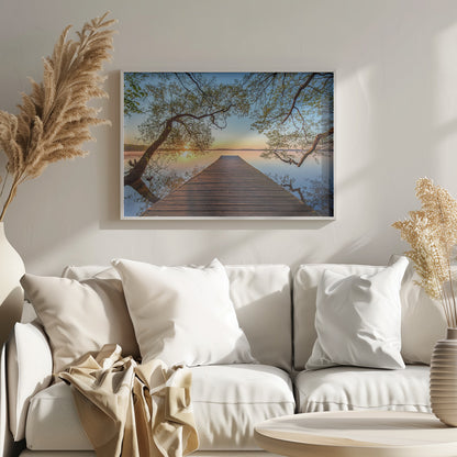 A tranquil sunrise over a still lake, viewed from a long wooden pier that stretches into the water, with silhouetted tree branches framing the serene scene. Poster