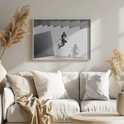 A framed, black and white photograph of a young person in mid-air, skillfully juggling a soccer ball with their foot against a clean, white brick wall. The person's shadow is cast sharply on the wall, mirroring the action, under a modern building with angled windows. Wall Art