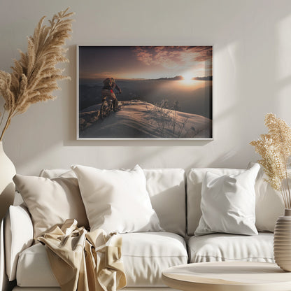 A mountain biker seen from behind pauses on a snowy mountain summit, overlooking a vast, misty mountain range as the sun sets, casting a warm orange and pink glow across the sky and snow. Artwork