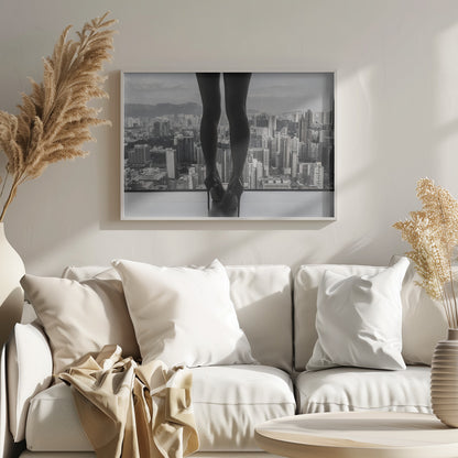 A framed black and white photograph showing the back of a woman's legs and high heels as she stands before a large window, looking out over a vast, dense cityscape with distant mountains under a cloudy sky. Artwork