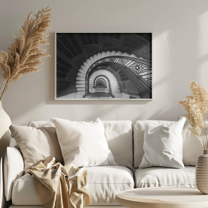 A mesmerizing black and white photograph shot from a high angle looking down into the center of a spiral staircase. The bright white marble steps contrast with the dark, ornate metal railing, creating a repeating arched pattern that draws the eye downward into a seemingly endless tunnel. The image is presented within a silver-colored frame. Artwork