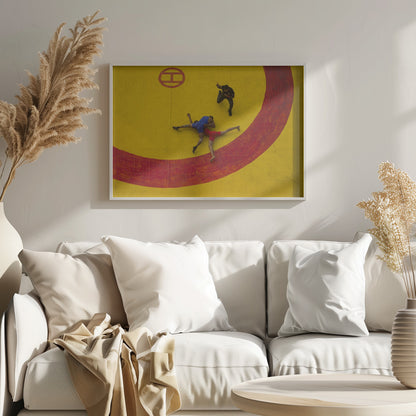 An aerial, high-angle photograph of a wrestling match in a silver frame. Two wrestlers, one in a blue singlet and the other in red, are locked in a hold on a vibrant yellow mat marked with a thick, painted red circle. A referee dressed in black stands nearby, gesturing. Artwork