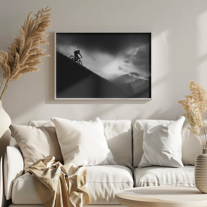A striking black and white photo of a mountain biker's silhouette on a steep decline, set against a dramatic, light-filled cloudy sky with distant, misty mountains. Artwork