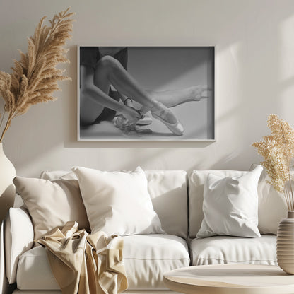 A black and white close-up photograph of a ballet dancer sitting on the floor, carefully tying the ribbons of a satin pointe shoe on her right foot. The image has a simple silver frame and captures a quiet moment of preparation. Poster