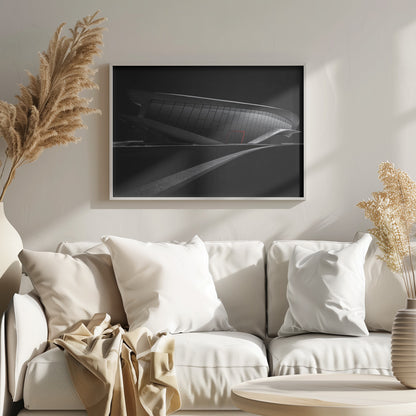 A dramatic black and white photograph of a futuristic, curved building with a dark, reflective facade. A single bright red door provides a striking pop of color. In the foreground, a light gray path curves across a dark landscape, leading the eye towards the structure. The entire image is presented within a silver-colored frame. Decor