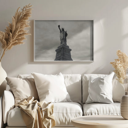 A low-angle, black and white photograph of the Statue of Liberty standing tall against a cloudy, dramatic sky, enclosed in a silver frame. Poster