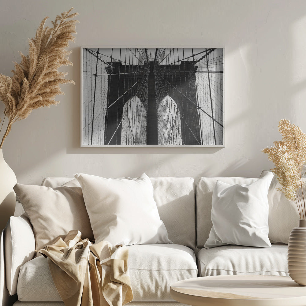A low-angle, black and white photograph looking up at the iconic stone towers of the Brooklyn Bridge. The intricate web of suspension cables and vertical suspender ropes creates a symmetrical, geometric pattern against the light sky. The photo is enclosed in a silver frame. Print