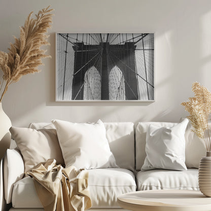 A low-angle, black and white photograph looking up at the iconic stone towers of the Brooklyn Bridge. The intricate web of suspension cables and vertical suspender ropes creates a symmetrical, geometric pattern against the light sky. The photo is enclosed in a silver frame. Print