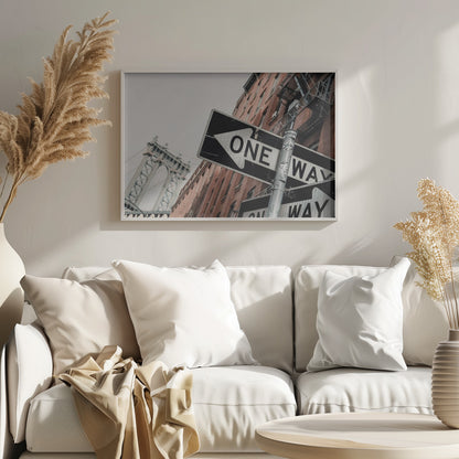 A low-angle shot of a black and white 'ONE WAY' street sign in the foreground, with a red brick building and the Manhattan Bridge visible in the background under an overcast sky, all enclosed in a silver frame. Artwork