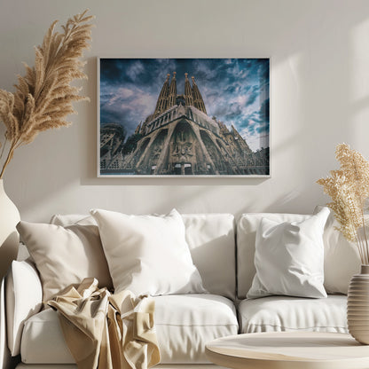 A dramatic, low-angle photograph of the Sagrada Familia in Barcelona, with its ornate stone facade and towering spires reaching up into a turbulent, dark blue and cloudy sky. The perspective emphasizes the grand scale and intricate detail of Gaudí's architecture. Print