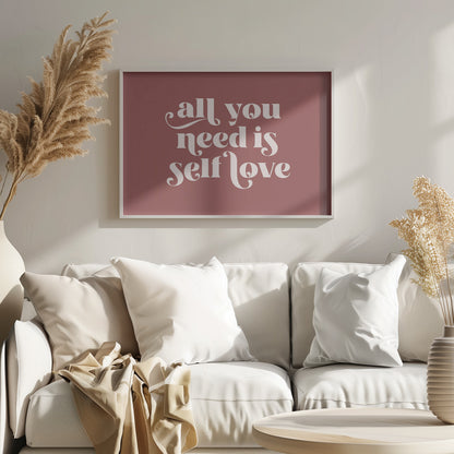 A framed art print with a dusty rose background. In the center, the phrase 'all you need is self love' is written in a creamy white, stylized, retro font. The artwork is enclosed in a simple silver frame. Decor