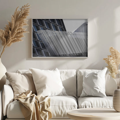 An abstract architectural photograph, framed in silver, showing a dynamic contrast between two modern building facades. In the background, a dark building features a grid of diamond-shaped windows with white frames, while a structure in the foreground creates a wave of fine, parallel white vertical lines against the sky and the other building. Print