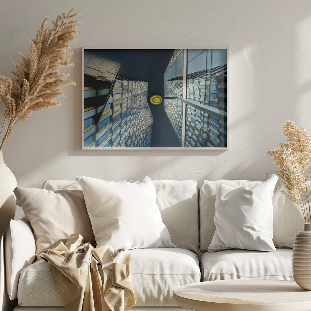A worm's-eye view looking up between two modern glass skyscrapers with geometric blue, yellow, and silver panels, creating a symmetrical reflection against a dark blue sky, with a bright yellow streetlamp centered in the gap. Decor