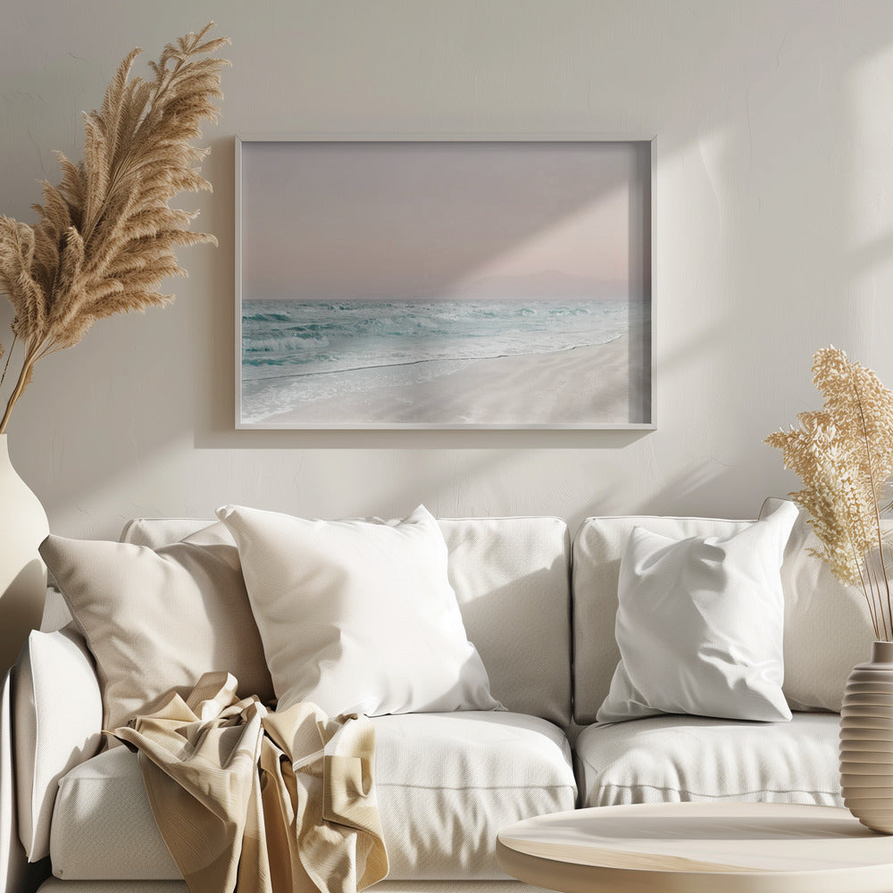 A serene coastal scene with gentle turquoise waves washing onto a sandy beach. The sky is a soft, hazy pink, and faint outlines of mountains are visible in the distant background. The artwork is presented in a silver frame. Print