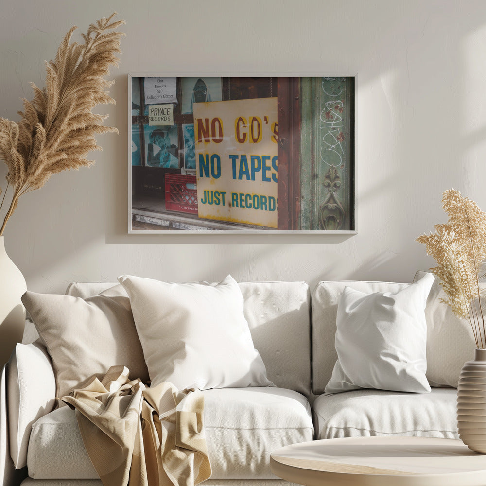 A weathered yellow sign in a record store window reads 'NO CD'S NO TAPES JUST RECORDS' in bold lettering. Behind the sign, vintage album covers and other posters are visible in the shop's display, next to a rustic green and brown door frame. Decor