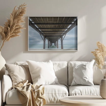 A symmetrical, low-angle photograph taken from underneath a long pier, looking out to sea. The rusty metal pilings and wooden planks of the pier create a receding perspective over the smooth, long-exposure water, all under a cloudy sky and presented within a silver frame. Poster