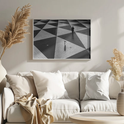 A high-angle, black and white photograph of a man in a coat and fedora walking away across a vast plaza with a geometric pattern of black and light gray triangles. An electric scooter stands alone in the foreground, creating a sense of solitude and contrast. The image is presented within a silver frame. Decor