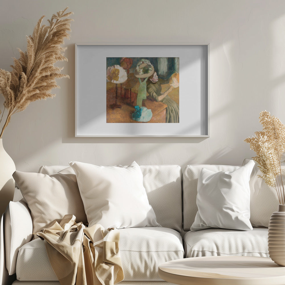 A framed print of Edgar Degas's Impressionist painting 'The Millinery Shop,' showing a woman in a green dress examining a hat in a shop filled with various fashionable hats on display stands. Print