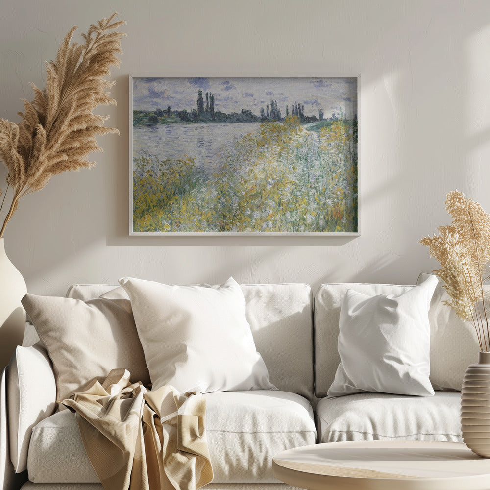An impressionist landscape painting of a riverbank covered in yellow and white wildflowers. A calm river reflects the cloudy sky, and the opposite bank is lined with tall, dark poplar trees. The artwork is displayed in a silver frame. Artwork