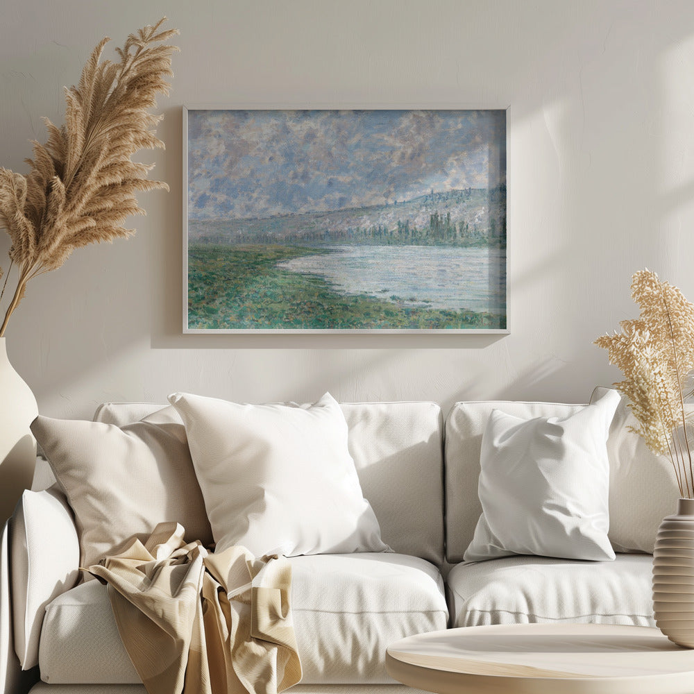 An Impressionist painting by Claude Monet, 'The Seine at Vétheuil', depicting a tranquil landscape. In the foreground, a lush green riverbank meets the calm, reflective water. A distant, soft-focus hillside rises under a vast sky filled with puffy white and grey clouds against a light blue background. The artwork is characterized by visible, textured brushstrokes and is displayed in a modern silver frame. Artwork