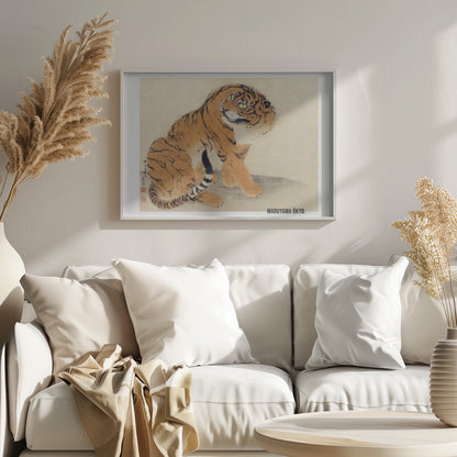 A vintage Japanese painting by Maruyama Ōkyo featuring a fluffy tiger sitting down and looking over its shoulder. The tiger has vibrant orange fur with black stripes and a white ruff. The background is a simple, textured beige paper with Japanese characters on the left. The artist's name is printed in the bottom right corner, and the entire piece is displayed in a silver frame. Print