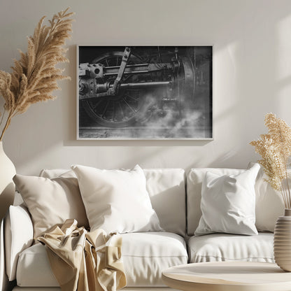 A dramatic, black and white close-up photograph of the wheels and driving rods of a steam locomotive. Puffs of steam rise from the tracks, partially obscuring the lower part of the machinery. Print