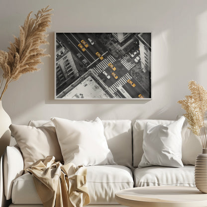 An aerial, black and white photograph of a bustling New York City intersection, with the iconic yellow taxi cabs highlighted in a vibrant selective color. Pedestrians cross the street as cars and a bus navigate the busy roads between tall buildings. Wall Art