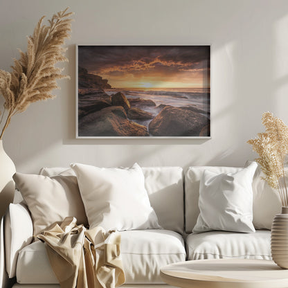 A framed photograph of a dramatic ocean sunset, with fiery orange clouds filling the sky. Large, dark rocks sit on the shore as long-exposure waves create a misty effect around them. Print