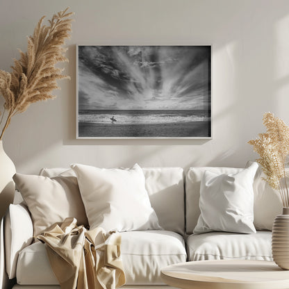 A dramatic black and white photograph of a lone surfer silhouetted against the sea, walking along a sandy beach. The sky is filled with spectacular, radiating clouds that stretch across the horizon, creating a powerful and moody atmosphere. The image is enclosed in a silver frame. Poster