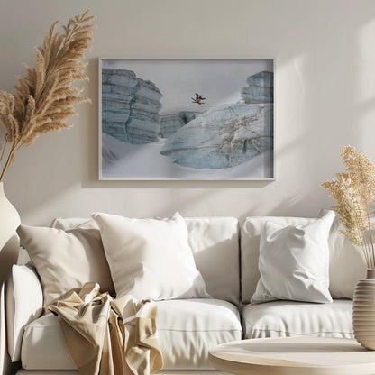 A framed photo of a skier in mid-air, jumping over a snowy crevasse between large, layered glacier formations under an overcast sky. The skier wears a black top and orange pants. Wall Art