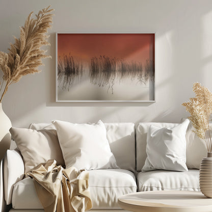 A minimalist landscape photograph in a silver frame, showing dark reeds silhouetted against a vibrant solid orange sky. The calm water perfectly reflects the reeds and the warm glow of the sky, creating a serene and abstract scene. Print