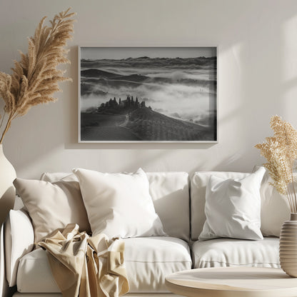 A framed, black and white photograph of a dramatic Tuscan landscape. A villa surrounded by cypress trees sits on a foreground hill, while thick fog blankets the rolling hills and valleys below. In the distance, a sea of clouds stretches towards a mountain range. Wall Art