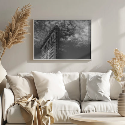A dramatic low-angle black and white photograph of the Flatiron Building in New York City, with its sharp, triangular corner pointing towards a sky filled with fluffy, white clouds. The image is framed with a brushed silver border. Print