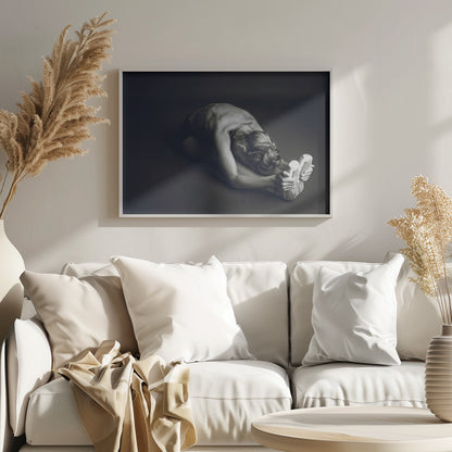 A black and white photograph of a ballerina stretching. She is seated on the floor in a deep forward fold, her torso bent over her legs as she holds onto her pointe shoes. The lighting accentuates the musculature of her back and shoulders, and her hair is styled in a braided bun. Decor