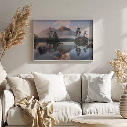 A serene mountain lake at sunrise or sunset, with a prominent peak illuminated by golden light. Small, rocky islands with pine trees dot the calm, clear water, which perfectly reflects the sky, trees, and mountains. A light mist hangs over the water and around the base of the mountains in the background. Decor