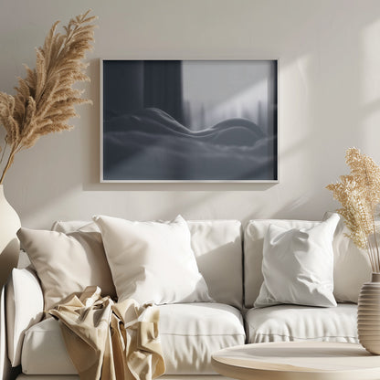 A close-up black and white artistic photograph capturing the soft curves of a person's back and hips as they lie down. The background is softly lit by a window, creating a play of light and shadow that emphasizes the human form's abstract, sculptural qualities. The image is displayed in a silver frame. Print