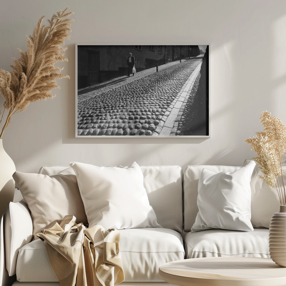 A framed, black and white photograph capturing an elderly man in a suit walking up a steep cobblestone street. The man carries a white bag and casts a long shadow behind him in the harsh sunlight. The street is lined with old, multi-story buildings, creating a classic European city scene. Poster