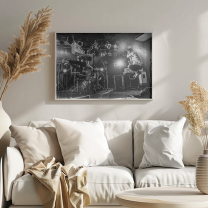 A high-energy, black and white photograph of a rock band performing live. The bassist and guitarist are frozen in mid-air as they jump, while the drummer plays in the background, all under bright stage lights. Print